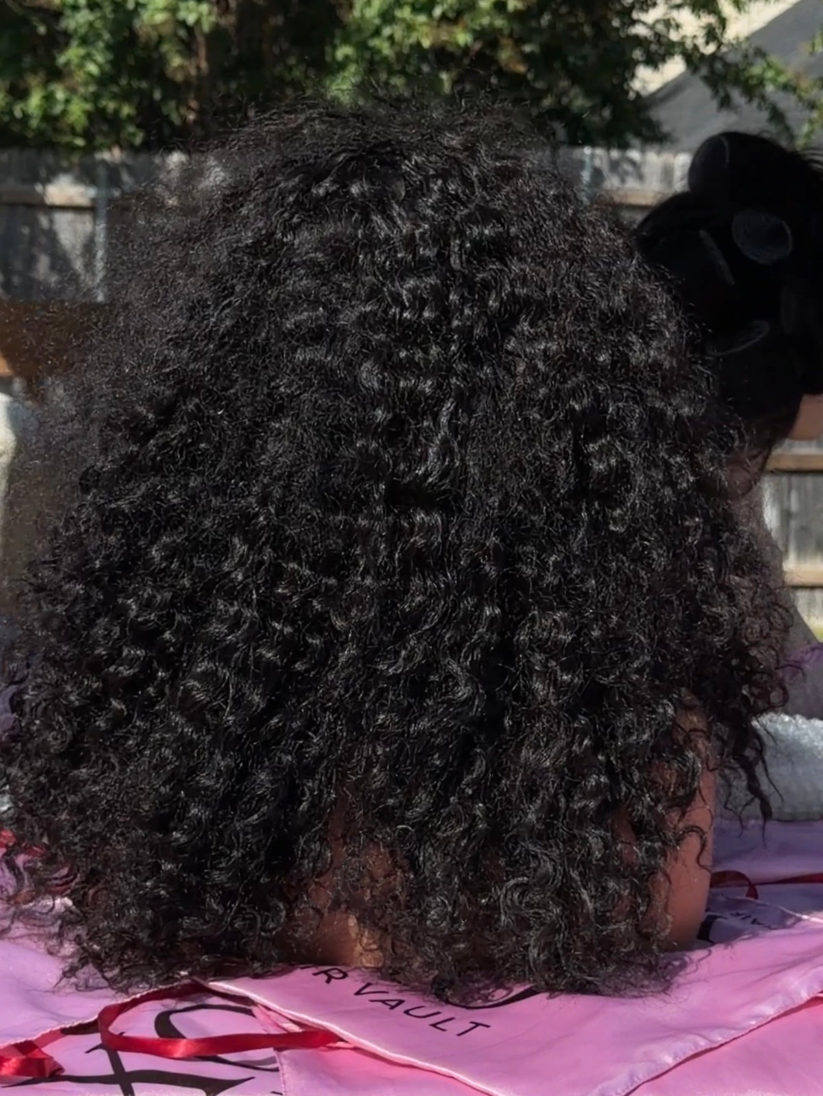 A close-up back view of a mannequin with voluminous, tightly curled black hair, wearing a black hair bow and sitting outdoors on a pink blanket with a wooden fence and greenery in the background.