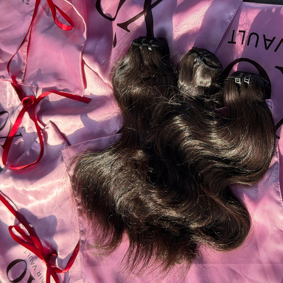 Three bundles of dark brown, wavy human hair extensions laid out on pink branded hair packaging with red ribbon ties.