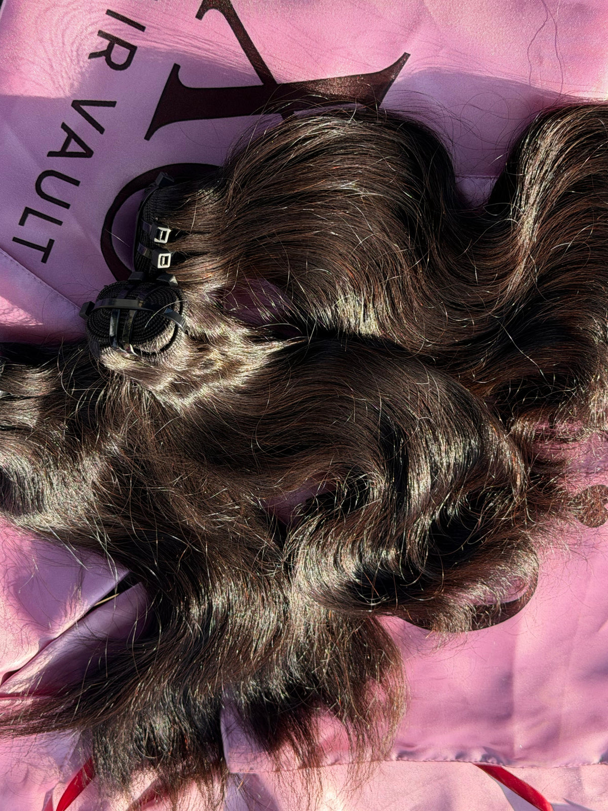 Three bundles of dark brown body wave hair extensions with black clip-in fasteners, laid out on a pink satin fabric with the brand name 'Hair Vault' printed on it.