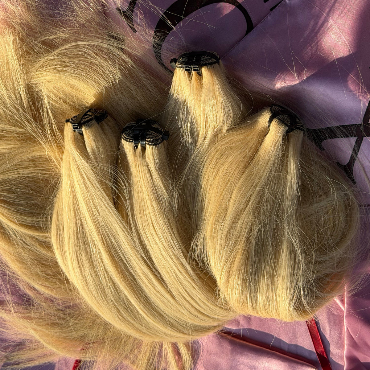 Three blonde hair wefts with black elastic ends, arranged on a pink hair salon bag with the brand name partially visible.