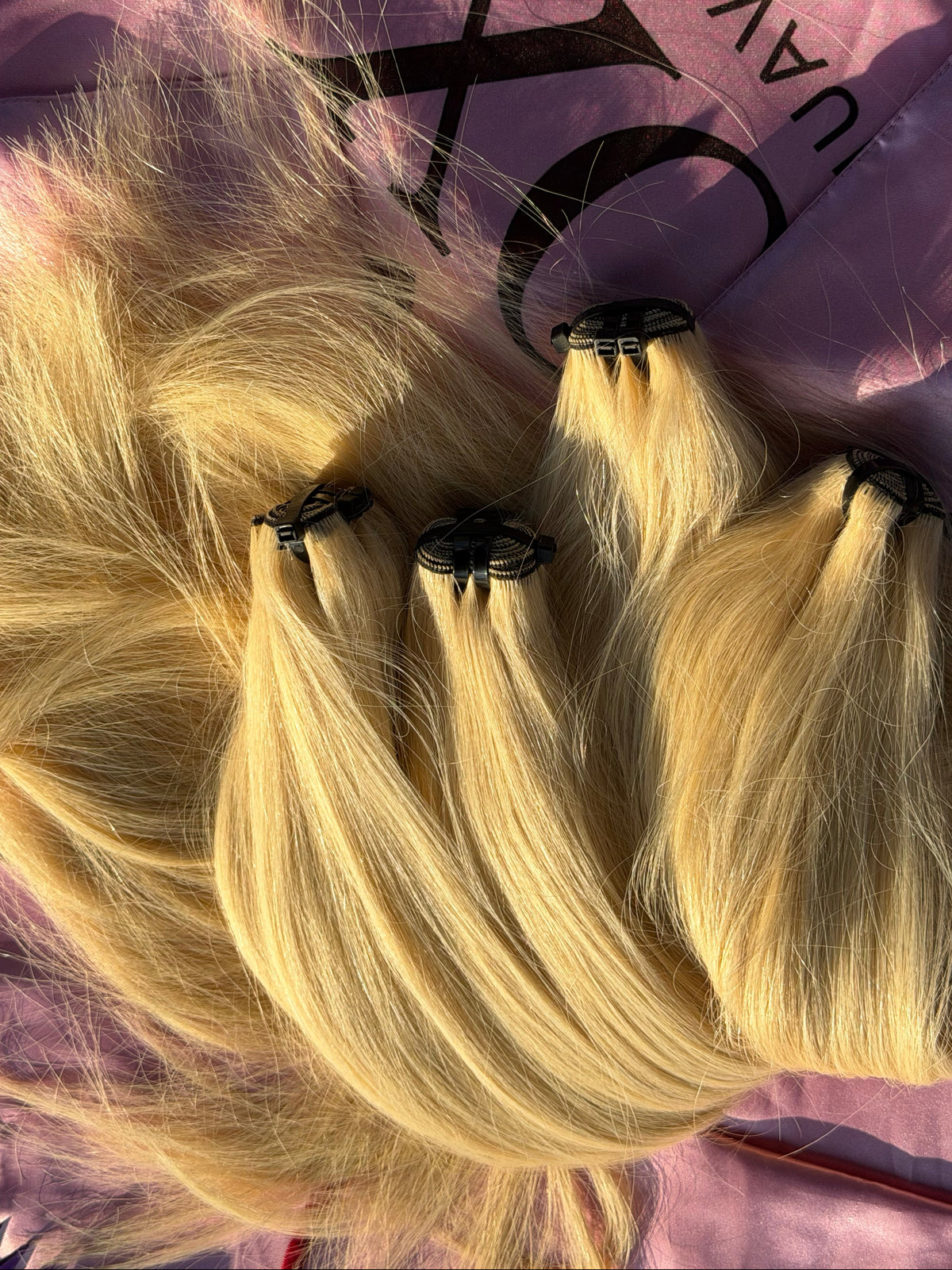 Four bundles of blonde hair extensions with black elastic ends, laid out on a pink branded hair bag.
