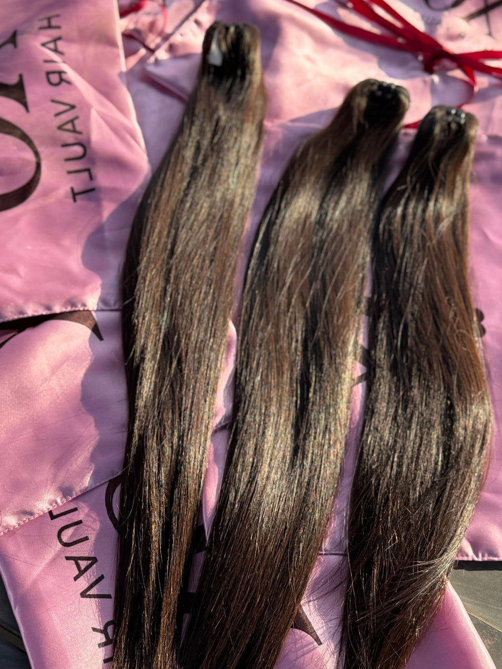 Three bundles of long, straight dark brown hair extensions laid out on pink satin hair bags with black text, photographed in bright natural sunlight.