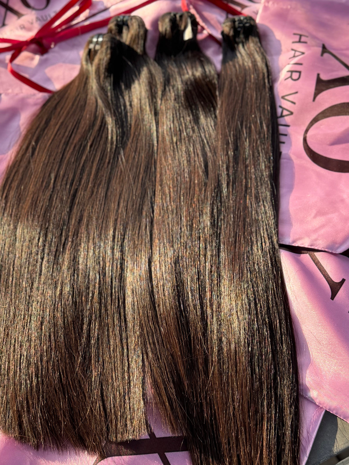 Three bundles of straight, dark brown hair extensions laid flat on a pink satin bag with printed text and red ribbon ties.