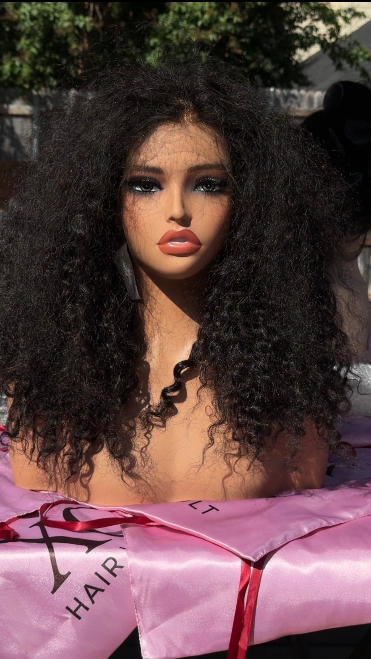 A close-up of a person outdoors wearing a voluminous, curly black wig. The person is wrapped in a pink satin cloth with a red ribbon, and the words 'HAIR' are printed on the fabric.