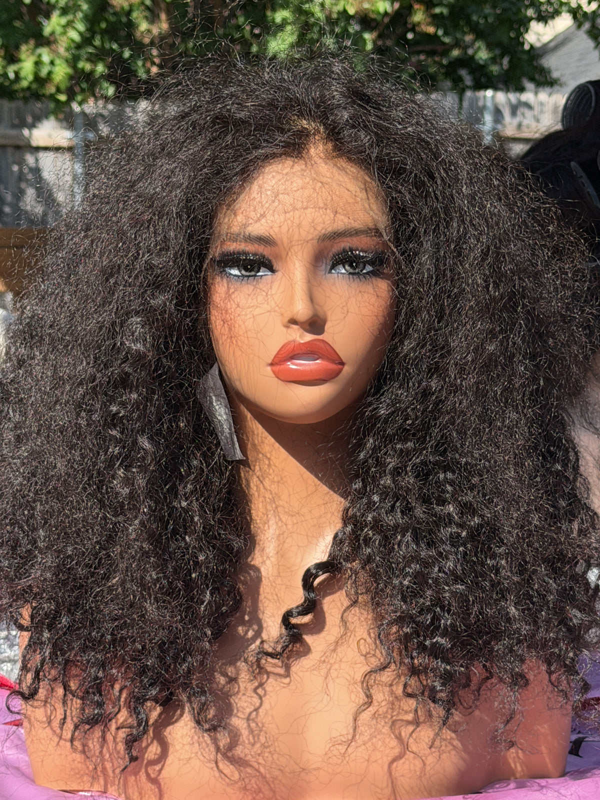 A close-up of a mannequin or doll head wearing a very voluminous, curly wig with dark brown to black coils. The wig is the product being shown, and because wigs are an exception where hair color counts as the product color, the color identity is based on the hair itself.