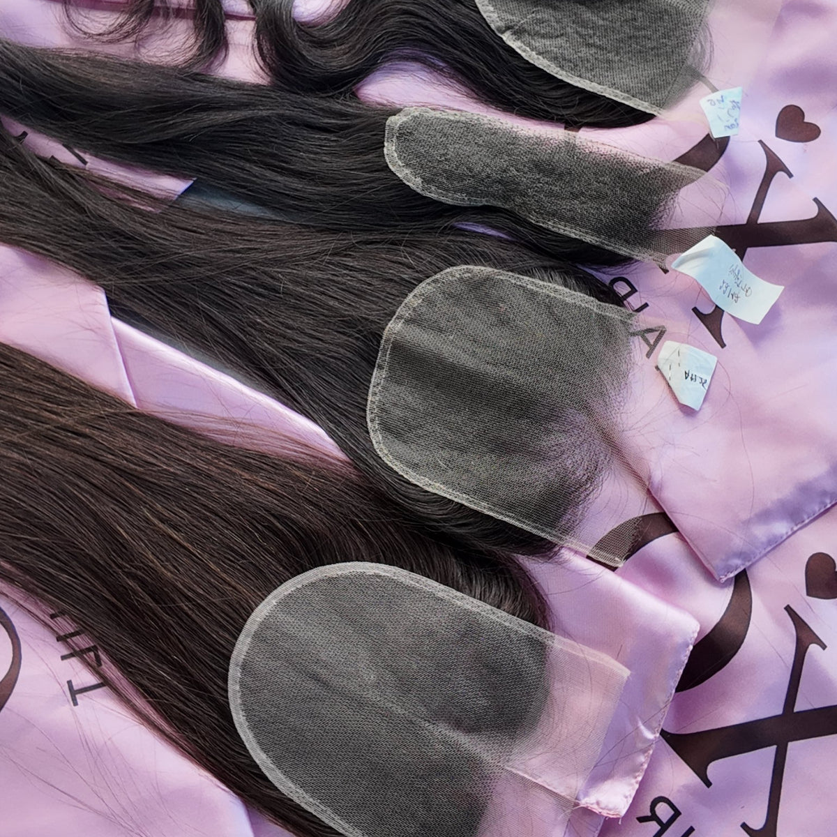 Four bundles of dark brown to black hair extensions laid out on pink satin hair bags with the XO Hair logo, photographed on a gray wooden surface.