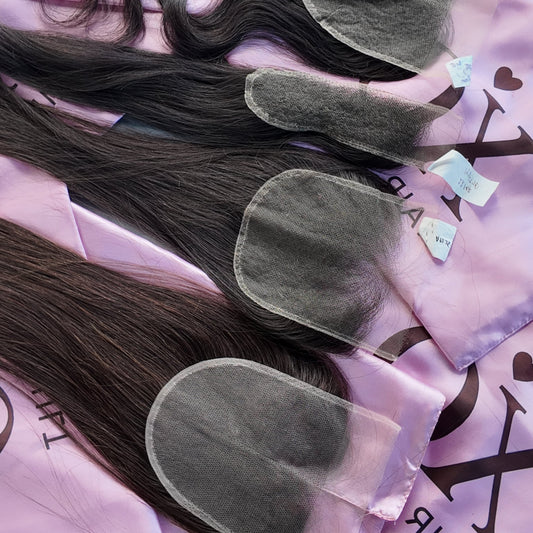 Four bundles of dark brown to black hair extensions laid out on pink satin hair bags with the XO Hair logo, photographed on a gray wooden surface.