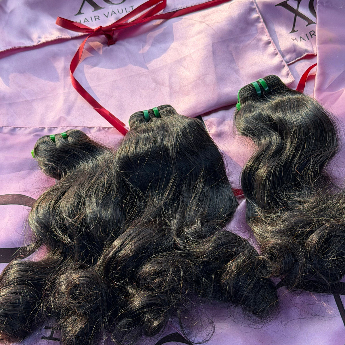 Three bundles of dark wavy human hair extensions laid on a shiny light purple XO Hair Vault branded bag with a red ribbon.