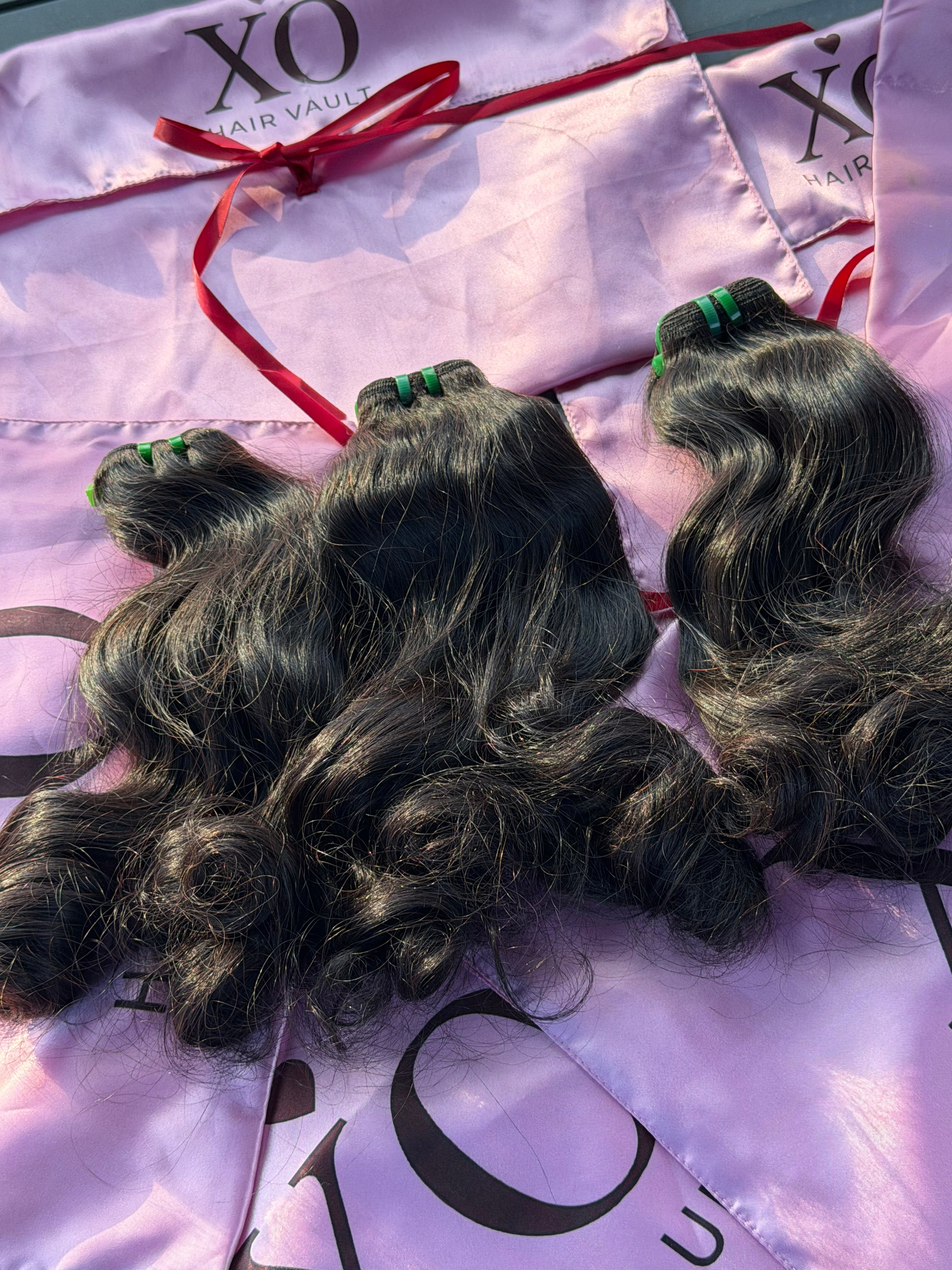Three bundles of dark wavy human hair extensions laid on a shiny light purple XO Hair Vault branded bag with a red ribbon.