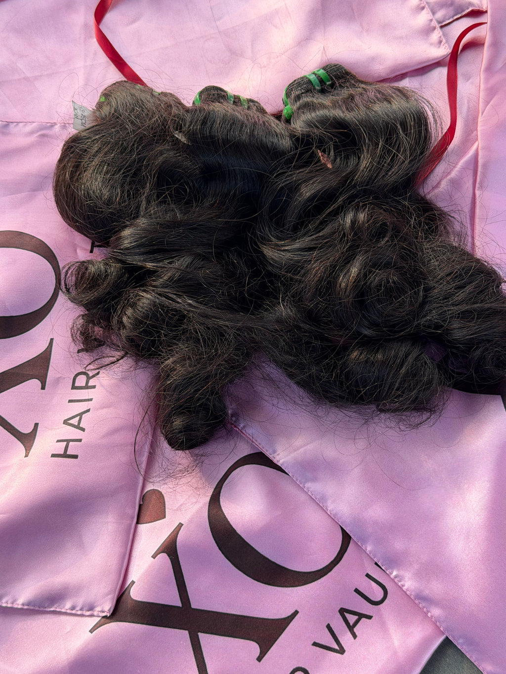 Three bundles of dark, wavy human hair extensions laid out on a pink satin cape with 'XO HAIR' branding.