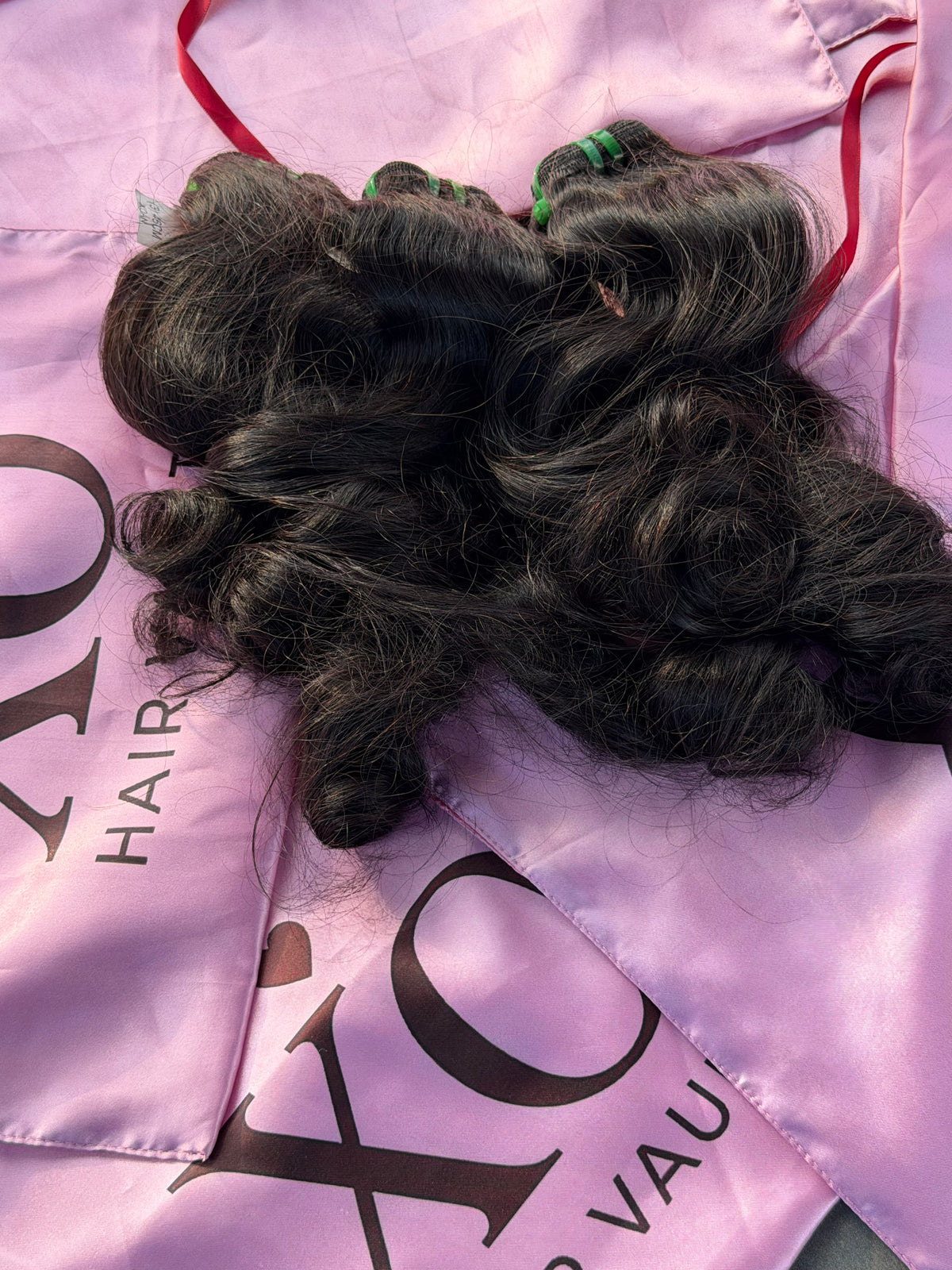 Three bundles of dark, wavy human hair extensions laid out on a pink satin cape with 'XO HAIR' branding.