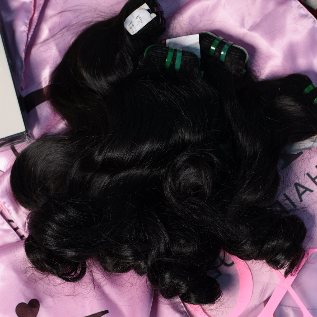 Three bundles of black wavy hair extensions laid out on a shiny pink satin background with 'XC HAIR VAULT' branding visible.