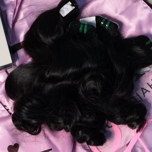 Three bundles of black wavy hair extensions laid out on a shiny pink satin background with 'XC HAIR VAULT' branding visible.