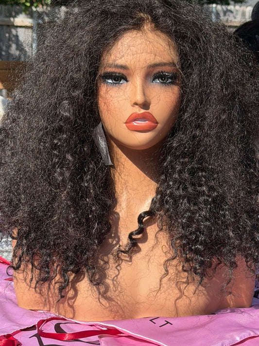 A close-up of a mannequin or doll head with a very large, voluminous dark curly wig, heavy makeup, and a pink garment at the bottom.