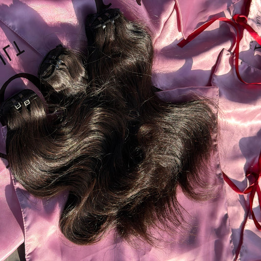 Three bundles of dark brown, wavy hair extensions laid out on a shiny pink satin fabric with red ribbon ties and black 'AIR VAULT' branding.