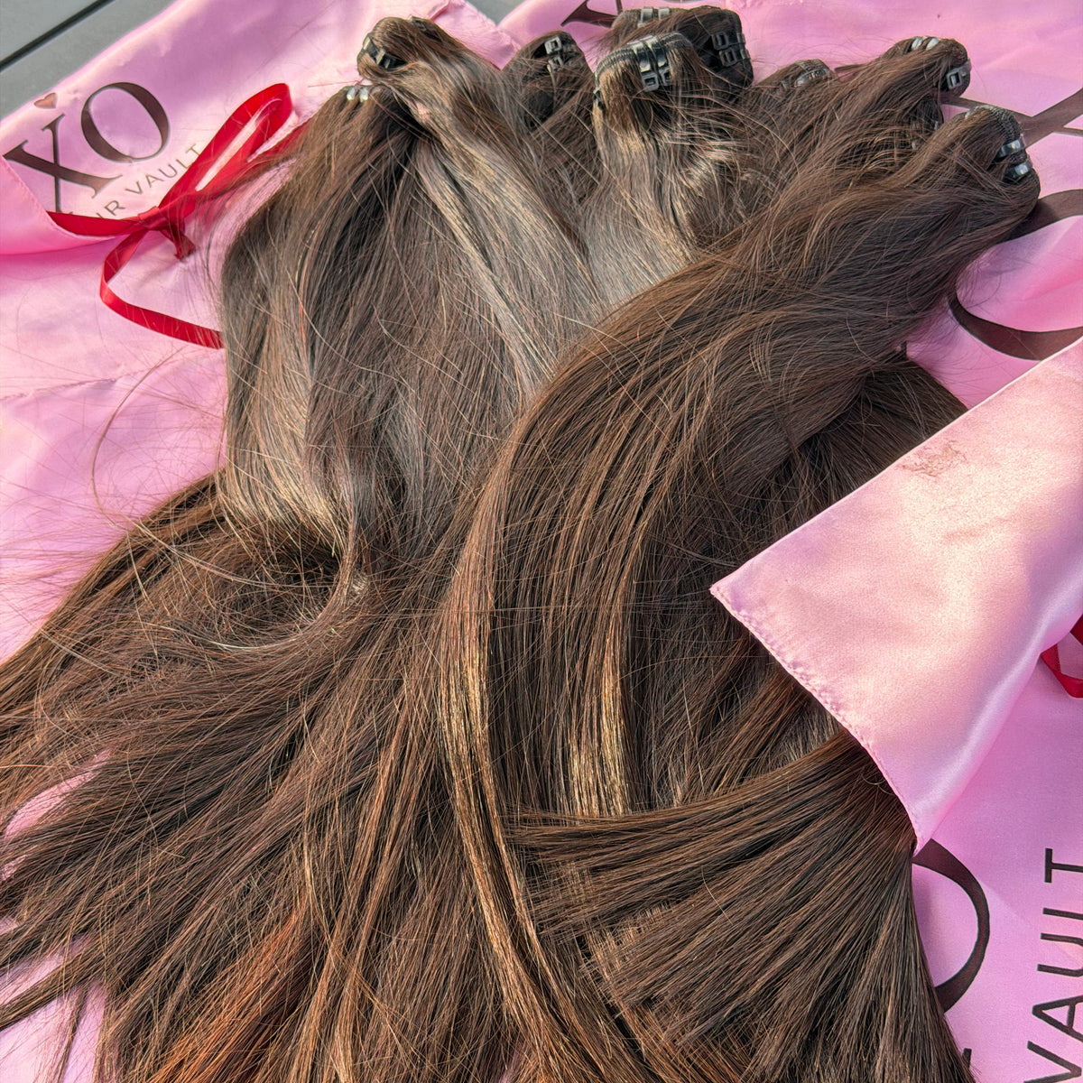 Three bundles of brown hair clip-in extensions with black adhesive tabs, laid out on pink branded hair extension packaging.